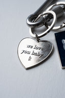 Silver metal keychain with polished finish, deep-blue tag engraved with Sleeves Studios icon, silver heart on back stamped “We love you, baby,” designed as a meaningful accessory