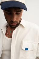 Man wearing a white shirt with a blue label, a navy cap, and a necklace with an emerald pendant.  - sleeves studios - white Button-up shirt - blue label - white shirt