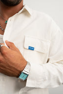 Person wearing a white shirt with a blue label, silver watch, and green necklace   - sleeves studios - white Button-up shirt - blue label - white shirt