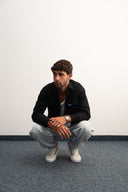 Man sitting on the floor against a white wall - sleeves studios - black Button-up shirt - blue label -  black shirt 