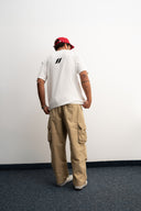 Person wearing a white t-shirt with a logo, beige cargo pants, and a red cap against a plain wall.