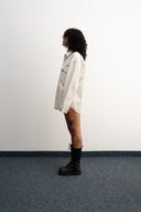 Person wearing a white oversized shirt and black boots standing against a plain wall  - sleeves studios - white Button-up shirt - blue label - white shirt