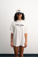 Person wearing a white oversized t-shirt with text and a cap on a plain background